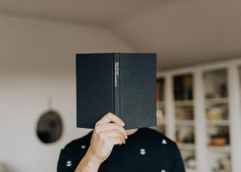 Faceless male holding opened black book in hand covering face while standing in middle of light room