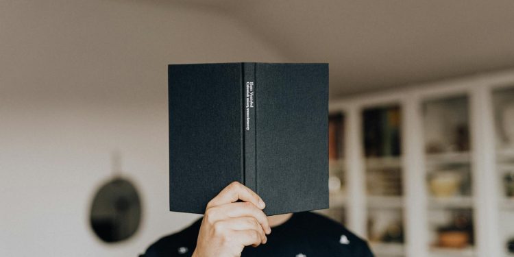 Faceless male holding opened black book in hand covering face while standing in middle of light room