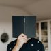 Faceless male holding opened black book in hand covering face while standing in middle of light room