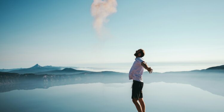 man standing on sand while spreading arms beside calm body of water